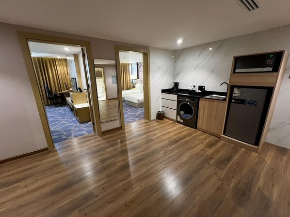 Una habitación de hotel moderna con suelo de madera, una zona de cocina con microondas y cafetera, y un dormitorio separado visible a través de una puerta abierta.
