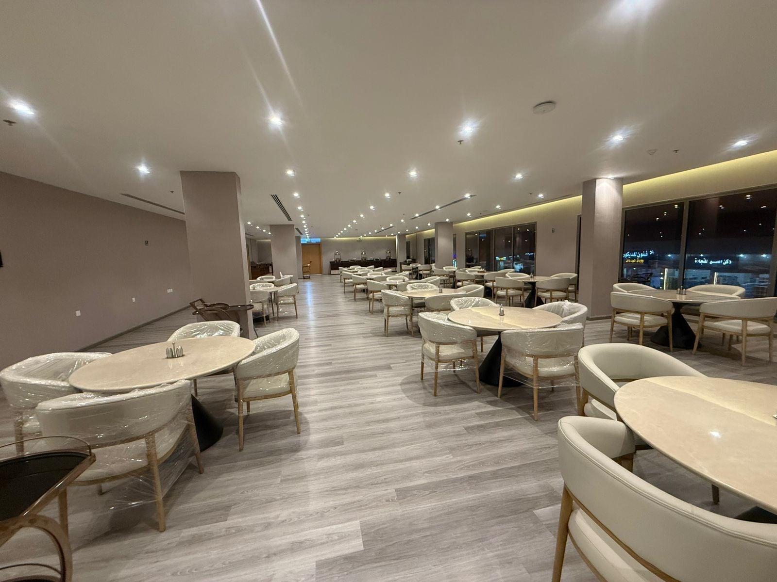 A modern dining area with round tables and chairs, a wooden floor, and a view of the city at night through large windows.
