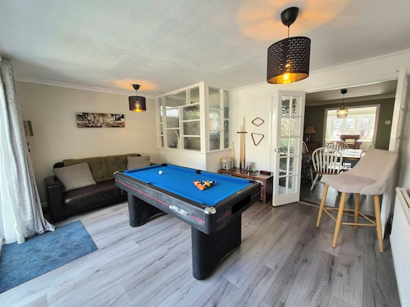 A pool table with a blue felt surface, a dark-colored pool cue, and a pool ball. There is a dark-colored hanging lamp above the table. The room has light-colored wooden flooring and a grey area rug.