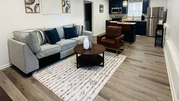 Un salon moderne avec un canapé gris, un fauteuil marron et une table basse. On aperçoit, au fond, une cuisine avec des meubles sombres et des appareils électroménagers en acier inoxydable.