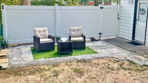 Un espace extérieur aménagé en terrasse, avec deux fauteuils en osier et une petite table, entouré d’un chemin de gravier et d’une zone de pelouse verdoyante.
