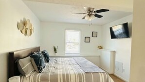 Une chambre à coucher comprenant un lit, une table de chevet, un ventilateur de plafond, une fenêtre et un téléviseur fixé au mur.