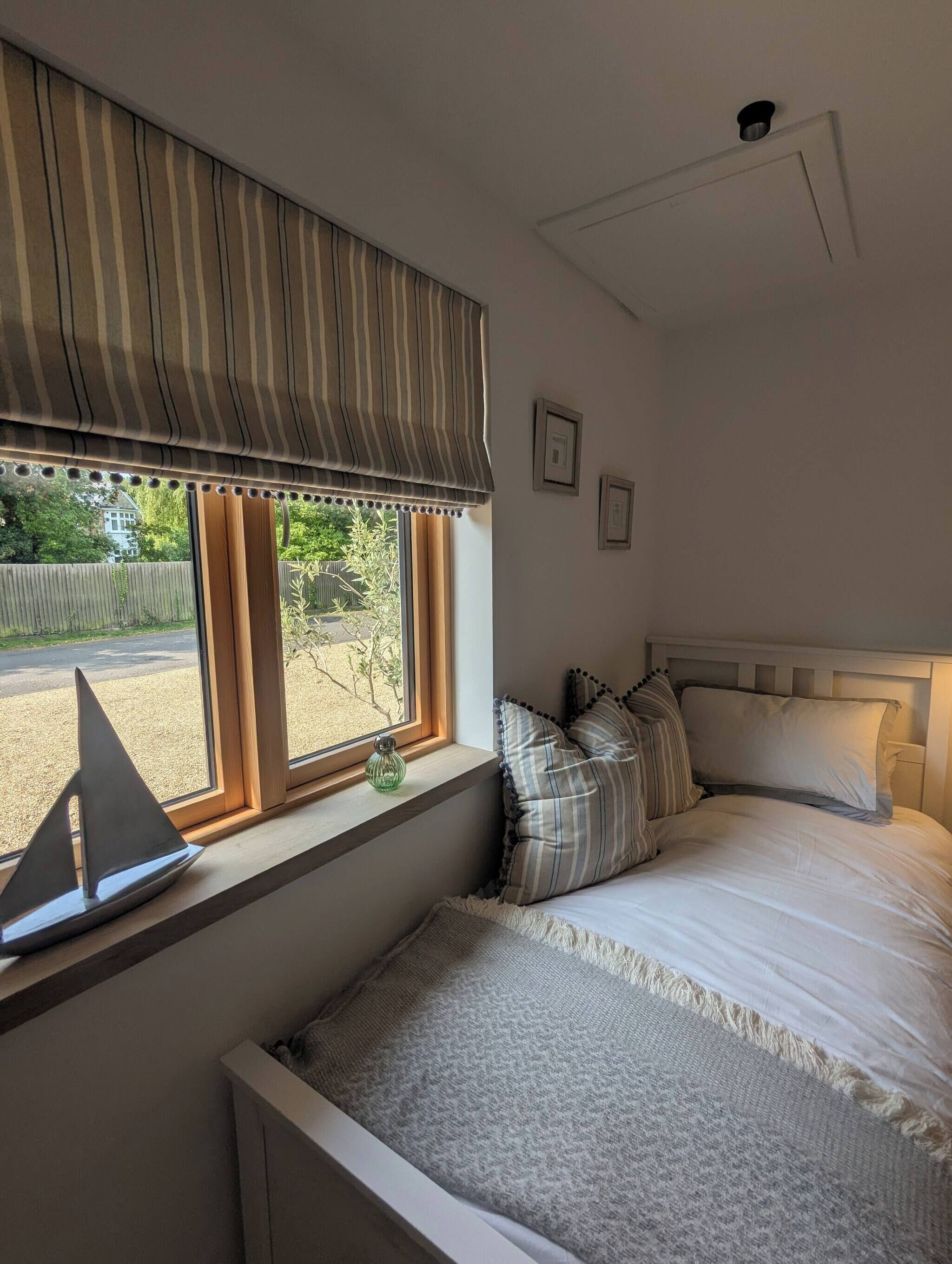 A bedroom with a bed, a window with a striped valance, a small table with a model ship, and a view of the outside.