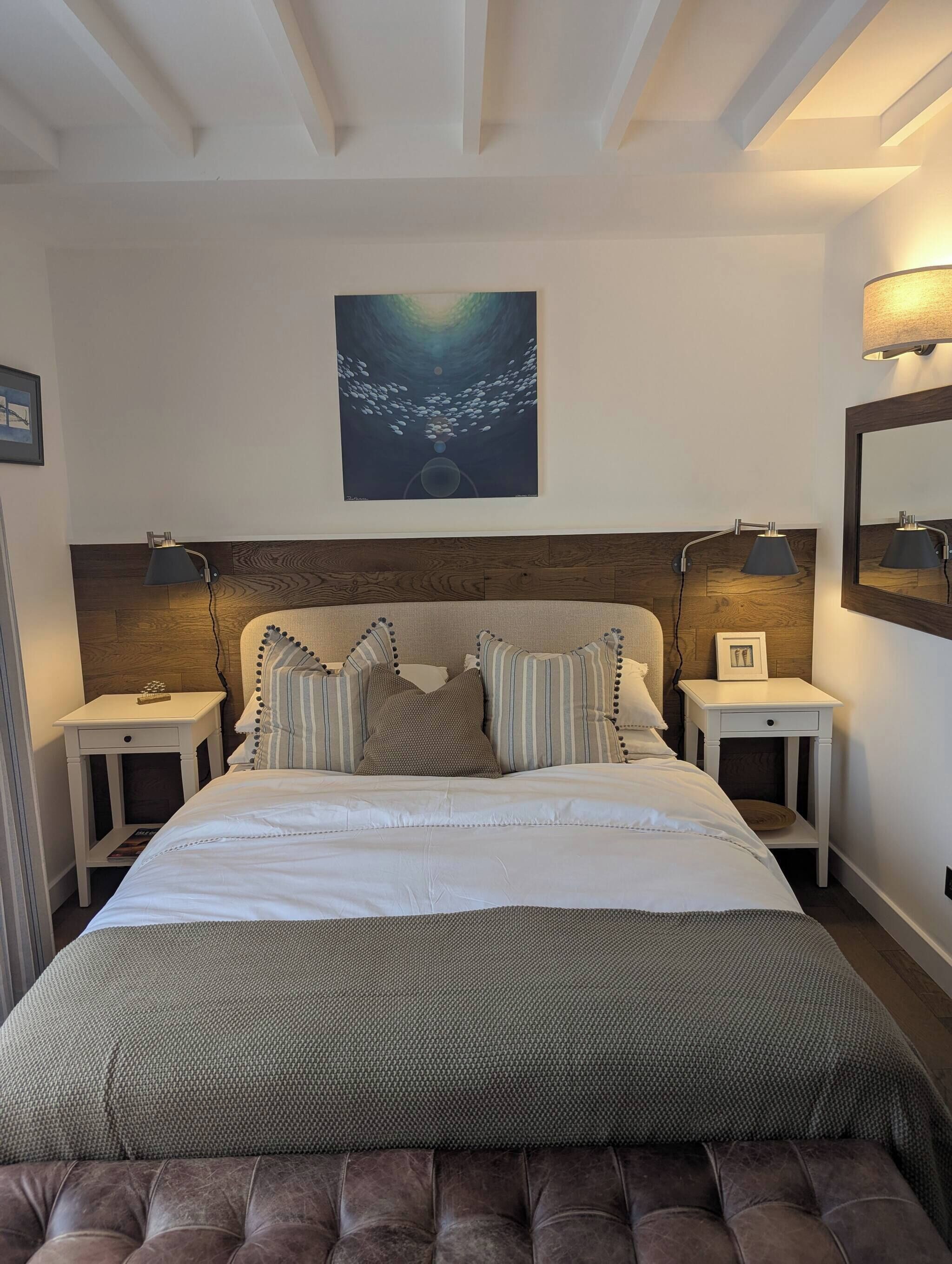 A bedroom with a large bed, two bedside tables with lamps, a headboard with a wooden panel, and a wall-mounted painting.
