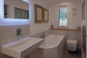 A modern bathroom with a freestanding bathtub, a vanity with a sink, a toilet, and a window with blinds. - Stylish New Apartment in Central Seaview - 5 Star Comfort! (Seaview)