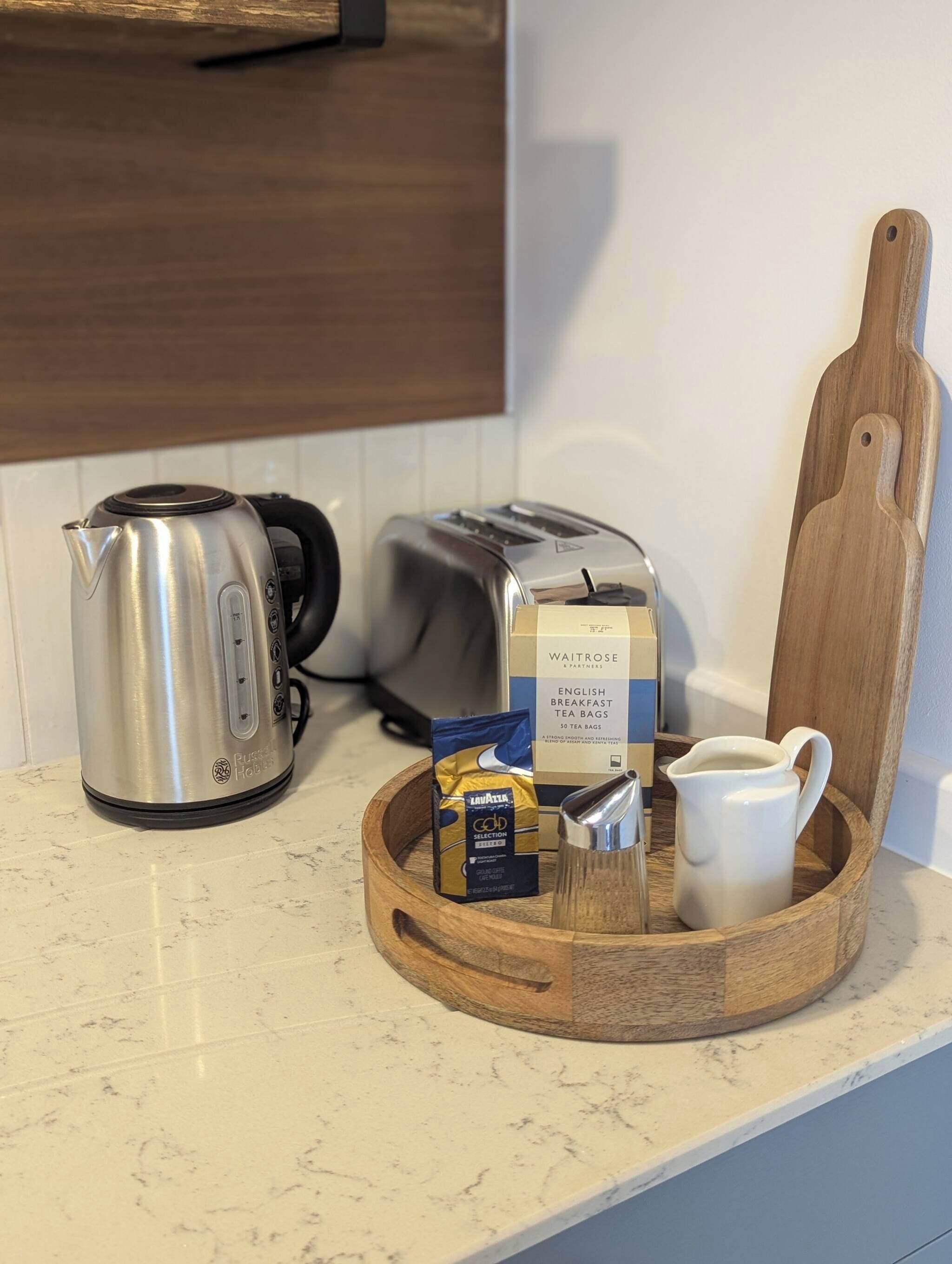 Una encimera de cocina con un hervidor, una tostadora, una tabla de cortar de madera, bolsitas de té y una jarra.