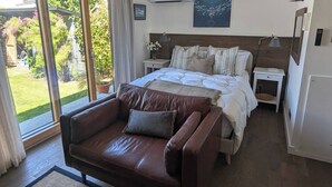 A bedroom with a large bed, a brown leather sofa, a nightstand, a lamp, and a view of the garden through a sliding glass door. - Stylish New Apartment in Central Seaview - 5 Star Comfort! (Seaview)