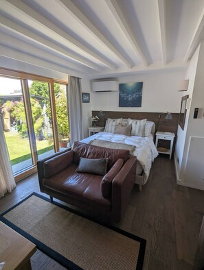 Ein Schlafzimmer mit einem großen Bett, einem braunen Ledersofa, einem Nachttisch, einer Lampe und Blick auf den Garten durch eine Schiebetür.