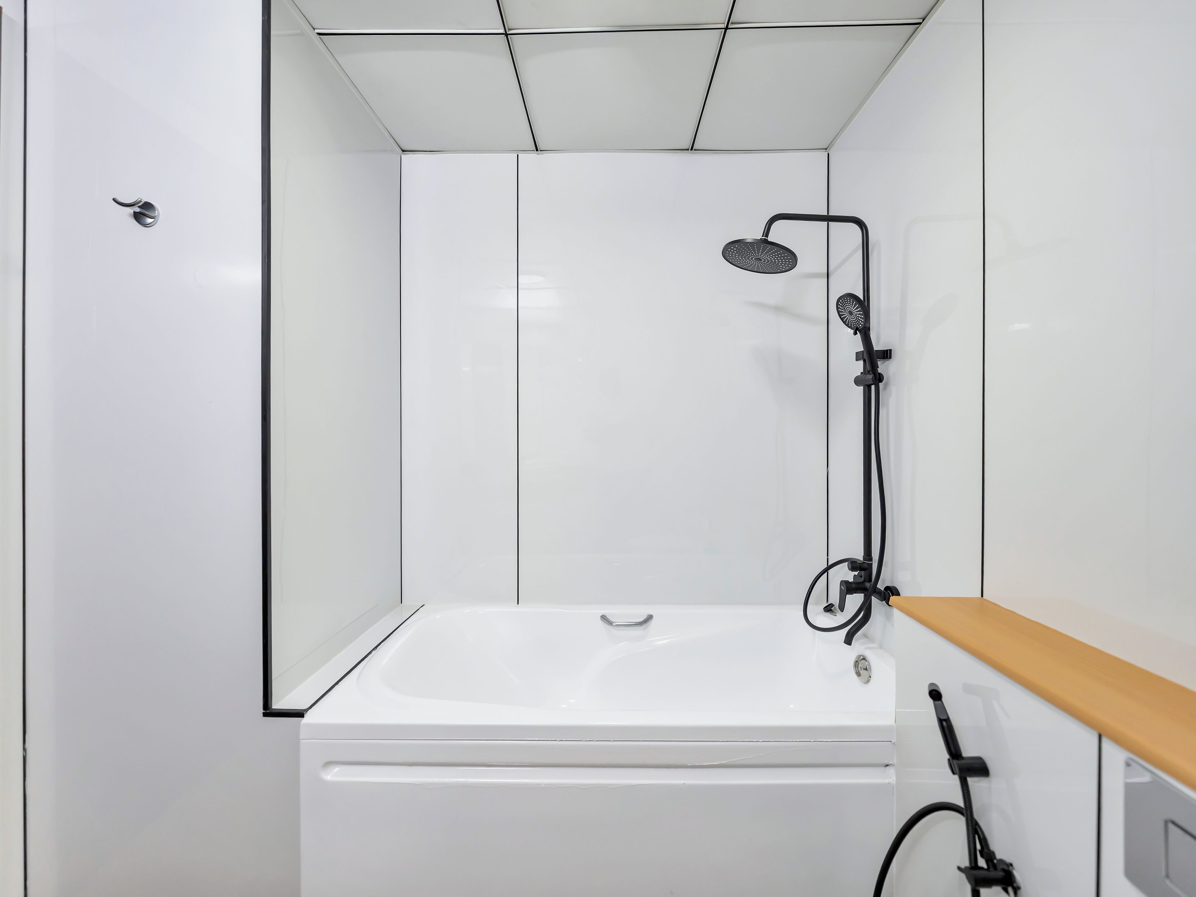 A modern bathroom with a white bathtub, a handheld showerhead, and a wooden shelf.