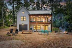 A two-story cabin with a balcony, a covered patio, and a fire pit area. - Eclectic Lakefront Cottage on Great East Lake! (Acton)