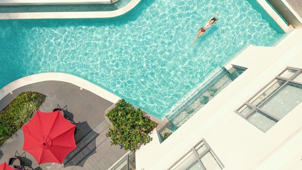 Eine Vogelperspektive zeigt einen Swimmingpool, in dem eine Person schwimmt, umgeben von einem Gebäude mit großen Fenstern und roten Sonnenschirmen.