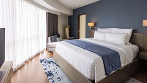 Ein modernes Hotelzimmer mit einem großen Bett, einem Nachttisch, einer Lampe und Blick ins Freie durch große Fenster.