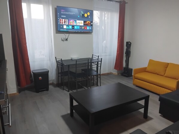 A living room with a TV mounted on the wall, a dining table with chairs, a sofa, and a vacuum cleaner. - Beautiful Brest Kérinou apartment refurbished (Brest)