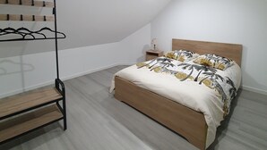 A bedroom with a sloped ceiling, a bed with a wooden headboard, a bedside table with a lamp, and a clothing rack with wooden shelves. - Gite des 3 têtes de loups (Fumay)