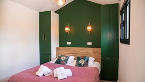 Uma cama bem arrumada com edredom rosa e duas almofadas, um guarda-roupa verde com duas luminárias, e uma janela com moldura preta.