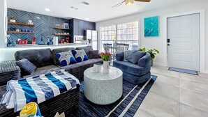A modern living room with a sofa, armchair, and round coffee table. There is a kitchen area with cabinets and a window.