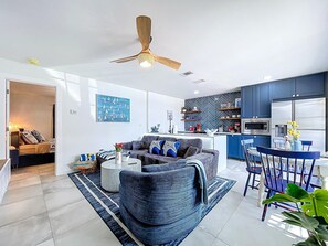 A modern living room with a sofa, coffee table, and a dining area with chairs and a table. There is a kitchen with blue cabinets and a refrigerator.