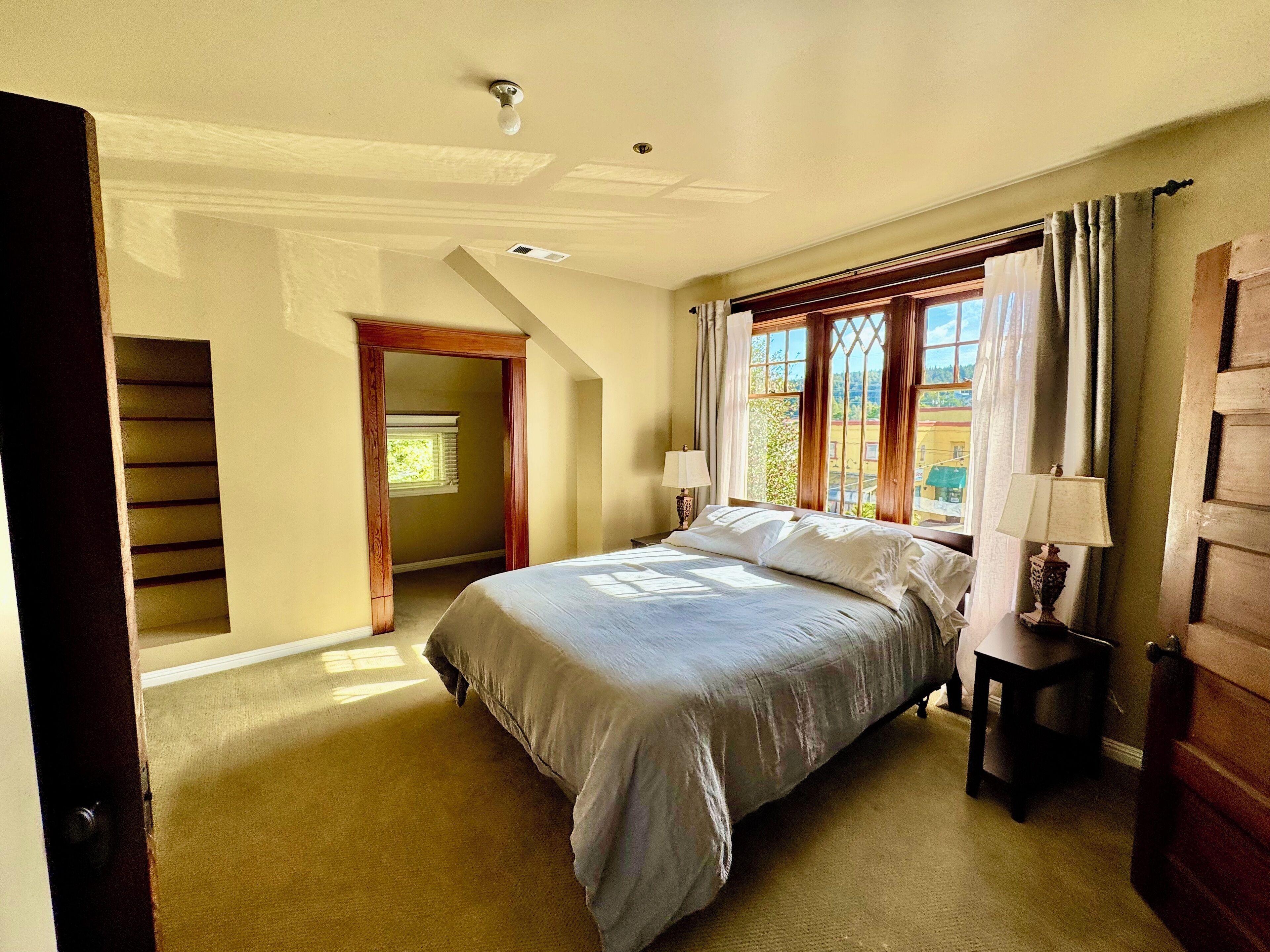 A bedroom with a bed, a nightstand, a window with curtains, a door, and a hallway leading to another room.