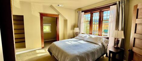 A bedroom with a bed, a nightstand, a window with curtains, a door, and a hallway leading to another room.