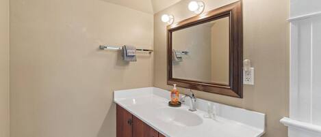 Un baño con un lavamanos blanco, un mueble de madera, un espejo con iluminación y un perchero para toallas.