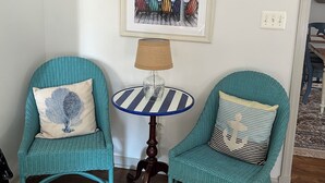 Twee smaragdgroene rieten stoelen met decoratieve kussens, een klein rond tafeltje met een gestreept blad, en een lijstwerk met een afbeelding van kleurrijke strandstoelen aan de muur.