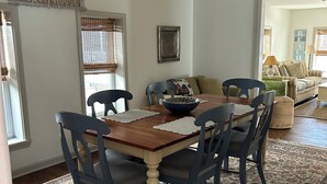 Een eetruimte met een houten tafel en blauwe stoelen, een kamerbreed tapijt en een uitzicht op een woonkamer met een bank en kussens.