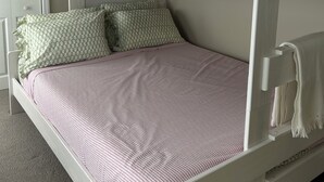 Een stapelbed met een wit frame, een onderste bed met een roze en witte geruit bedsprei, en een kussen met een groen patroon.