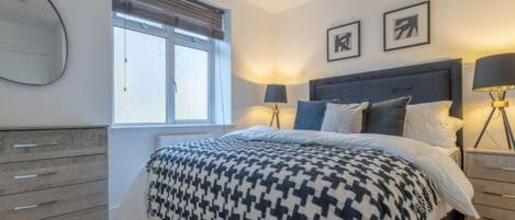Ein Schlafzimmer mit einem Bett, zwei Nachttischen, einer Kommode, einem Fenster mit Jalousien und zwei gerahmten Bildern an der Wand.