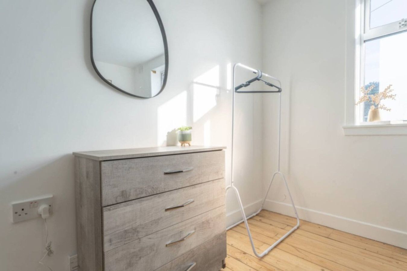 Une pièce comprenant une commode en bois, un miroir, un lampadaire et une fenêtre donnant sur le ciel.