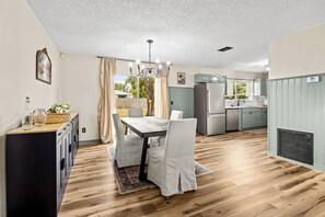 Una cocina moderna con comedor, que cuenta con piso de madera, sillas blancas y un candelabro.