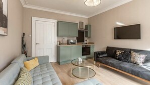 Un salon comprenant un canapé, une table basse, un divan en cuir noir et un coin cuisine avec des meubles verts et un four à micro-ondes.