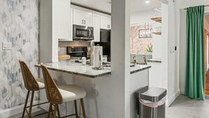 Eine moderne Küche mit weißen Schränken, einer Mikrowelle und einer Kaffeemaschine. Zwei Korbstühle und ein Mülleimer sind ebenfalls vorhanden.