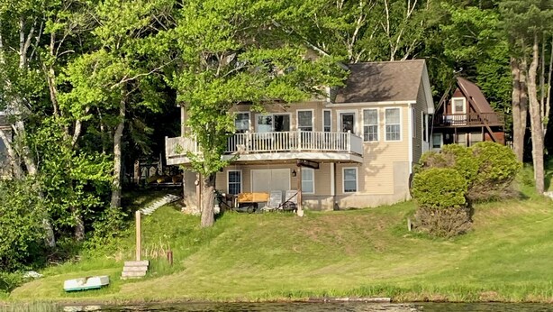 Uma casa de dois andares com varanda, situada perto de um lago rodeado por árvores.