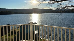 Um deck de madeira com guarda-corpo branco, com vista para um lago sob um céu claro e o sol brilhando.