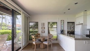 Una cocina moderna con zona de comedor, que incluye un gran ventanal con persianas, una mesa de madera con sillas y una encimera de cocina con electrodomésticos.
