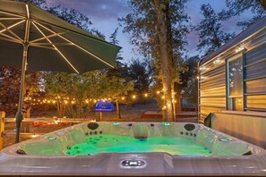 A hot tub with a control panel, surrounded by trees and string lights, with a covered area and a building in the background.