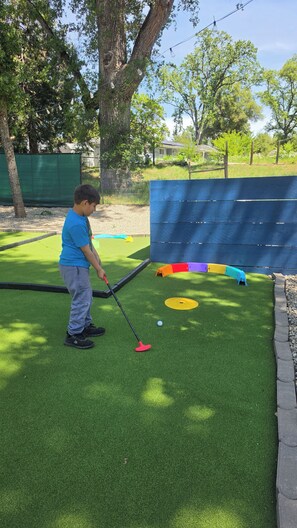A person playing mini-golf on artificial turf with a blue fence and trees in the background. - Dome Getaway, Hot Tub, Mini Golf, Game Room, Pets (Oakhurst)
