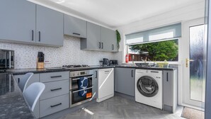 A modern kitchen with grey cabinets, a built-in oven, a washing machine, and a window with a view of greenery.