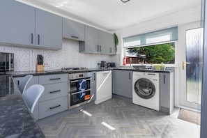 Une cuisine moderne avec des meubles gris, un four encastré, un lave-linge et une fenêtre donnant sur un espace verdoyant.