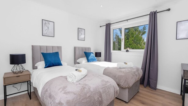 A hotel room with two beds, a window with curtains, a bedside table with a lamp, and artwork on the wall.