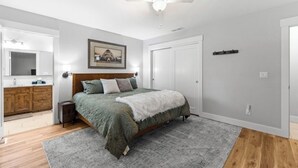 Un dormitorio con una cama, ventilador de techo, cómoda de madera, un cuadro enmarcado y una puerta.