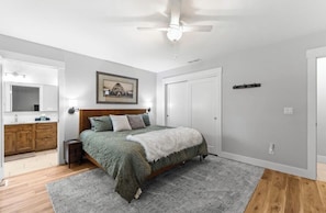 Un dormitorio con una cama, ventilador de techo, cómoda de madera, un cuadro enmarcado y una puerta.
