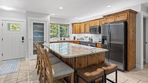 Cocina moderna con armarios de madera, una isla grande con taburetes de bar, electrodomésticos de acero inoxidable y vistas al exterior a través de amplios ventanales.