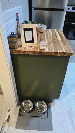 A kitchen counter with a stainless steel refrigerator, a microwave, and a trash can. There are two metal bowls on a mat below the counter.