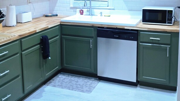 Una cocina con gabinetes verdes, encimera blanca, pileta, microondas y lavavajillas.