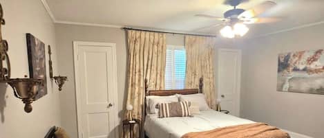 Ein Schlafzimmer mit Bett, Deckenventilator, Fenster mit Vorhängen, einer Tür und einem Bild an der Wand.