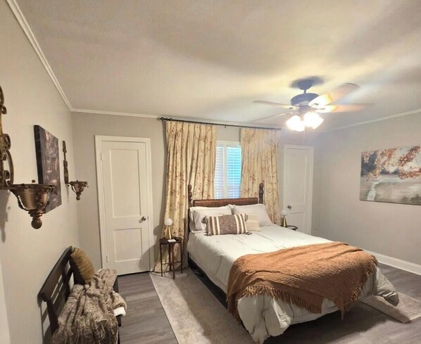 Un dormitorio con una cama, ventilador de techo, ventana con cortinas, una puerta y un cuadro en la pared.