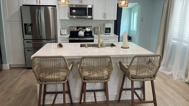 A modern kitchen with white cabinets, stainless steel appliances, a marble island, and wicker bar stools. - Delightful 2 bedroom - Island Cottage (Key Largo)