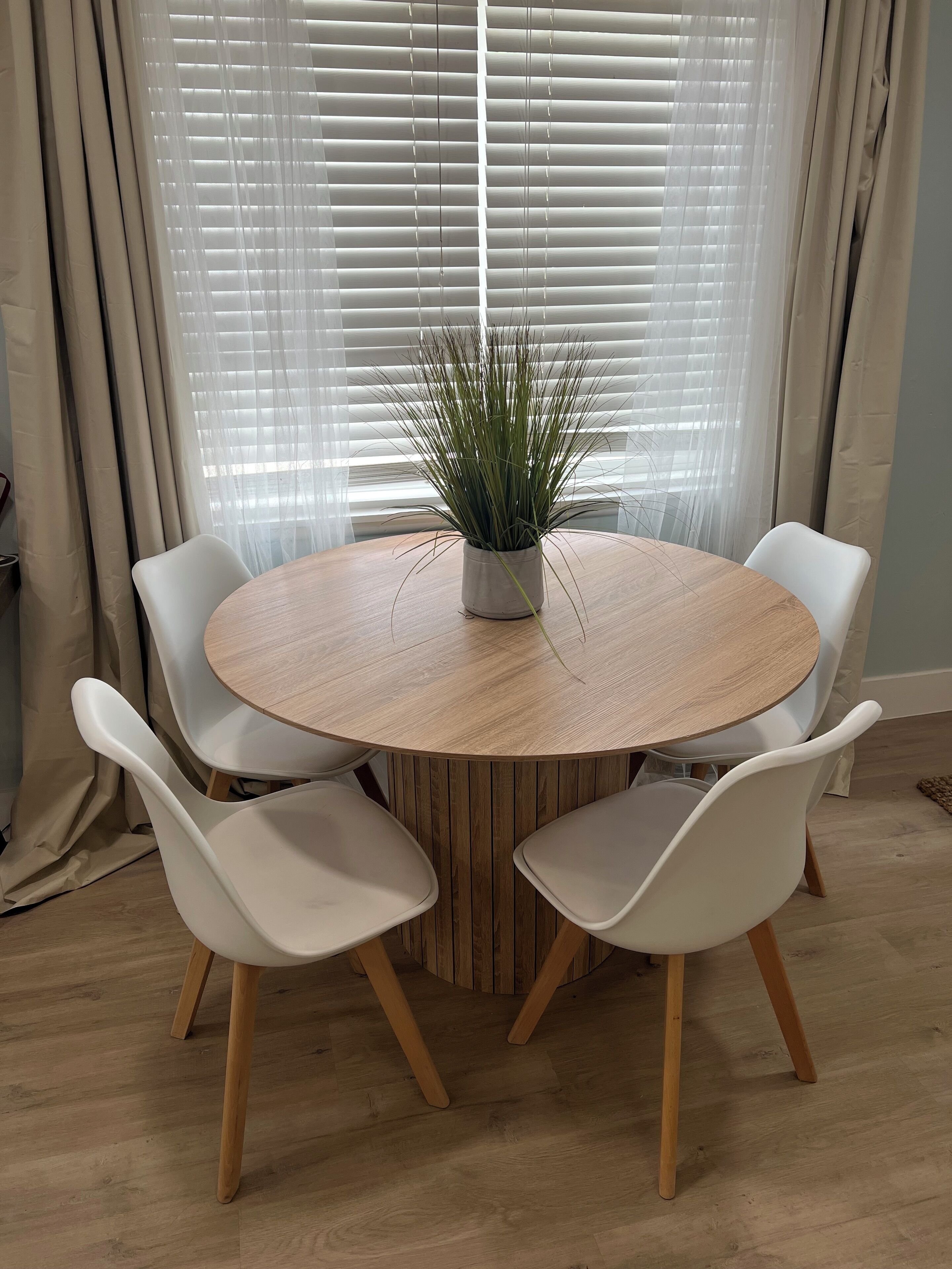 A round wooden table with four white chairs, a potted plant, and sheer white curtains.