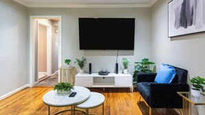 A modern living room with a flat-screen TV mounted on the wall, a white entertainment unit, a black sofa with a blue pillow, and a round coffee table with a remote control. - Culmore TX (Houston)